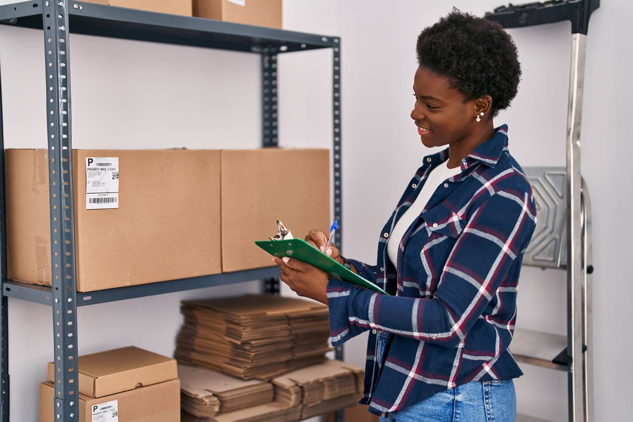 A woman holds a clipboard while standing next to boxes on a shelf. 