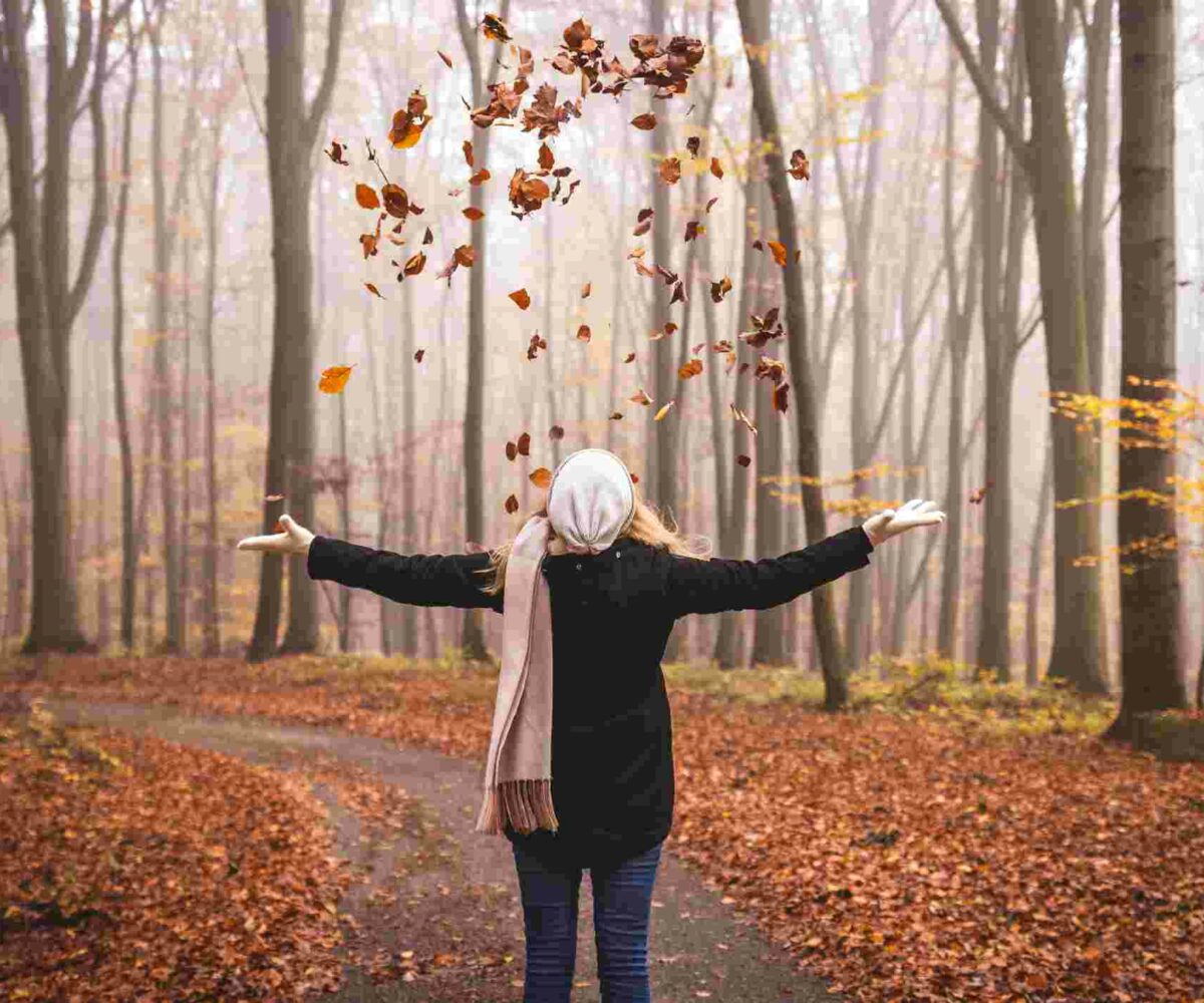 Woman celebrating the fall season sitting in a pile of leaves and throwing leaves into the air.