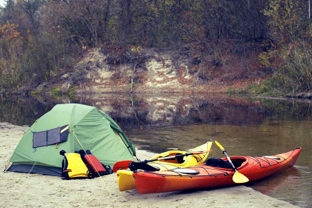 Two kayaks rest on the lakeside next to a tent and hiking packs.